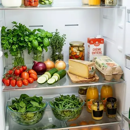 Un frigo ouvert avec des herbes, des tomates, une courgette, du comté, des œufs, de la crème et des bocaux